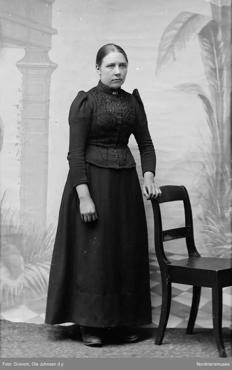 Portrett i helfigur av kvinne i kjole som holder i en stol. 