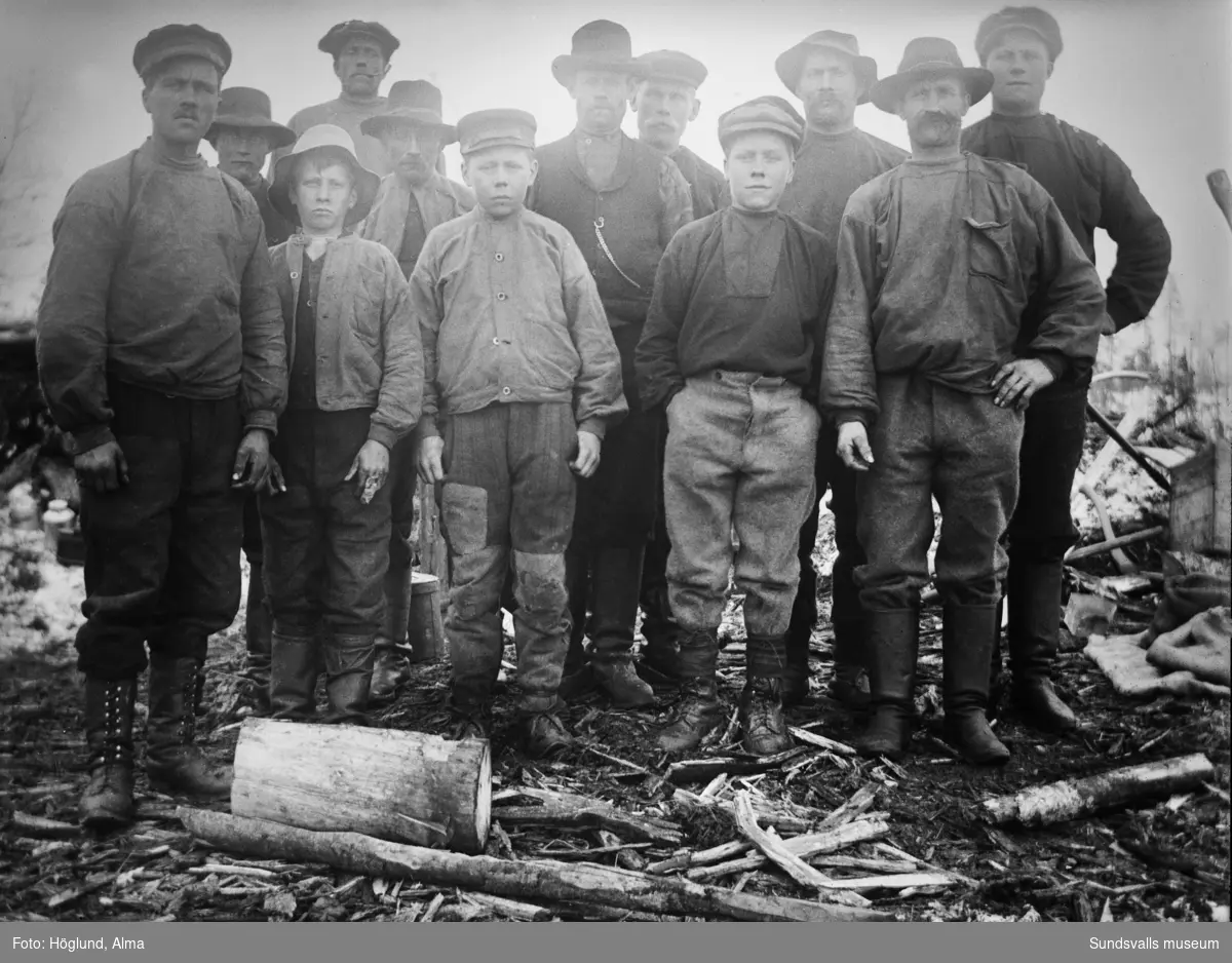 Gruppbild med arbetsklädda män och pojkar. Möjligen skogsarbetare.