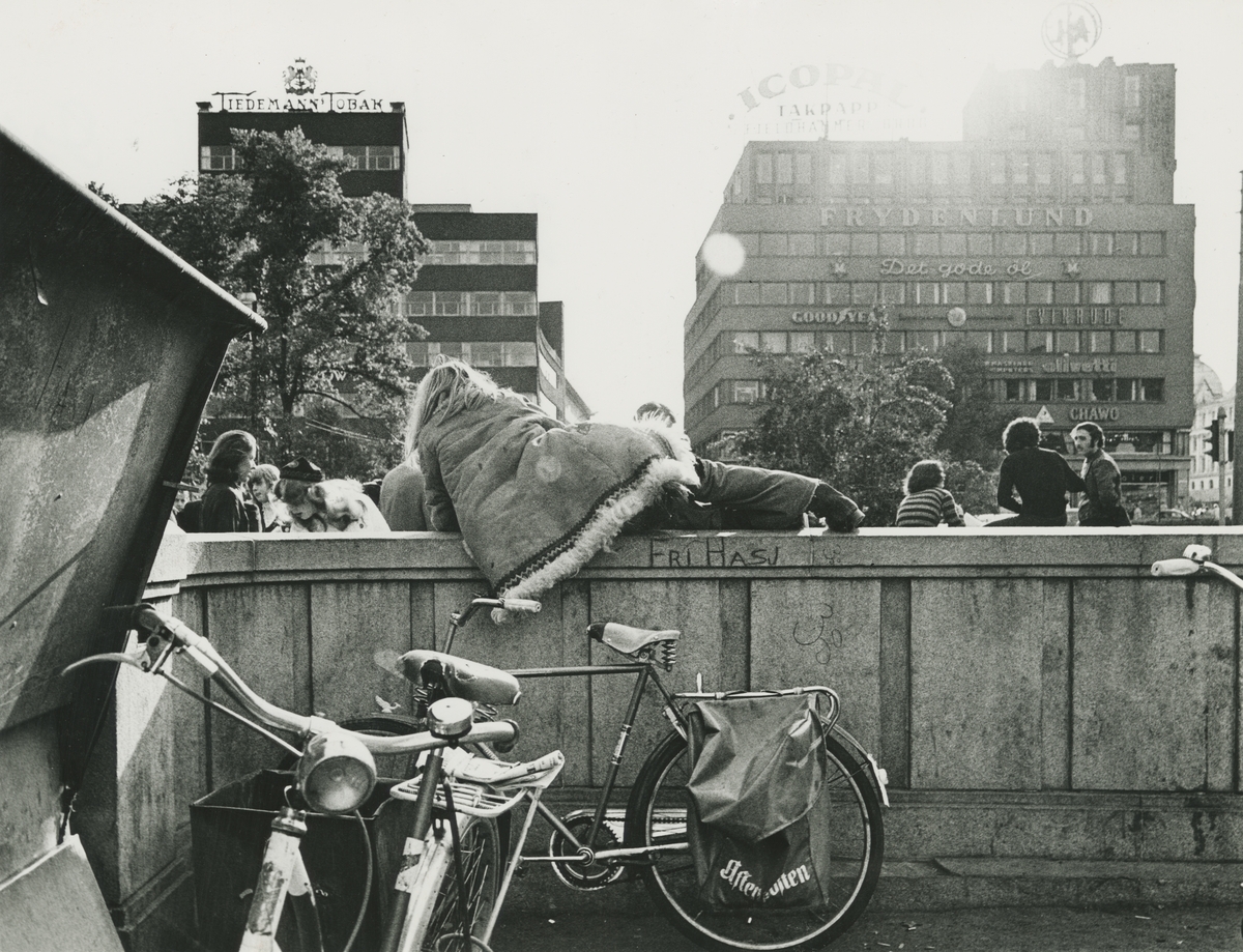 Ungdom og sykler ved nedgangen til Nationaltheateret stasjon. En person ikledd "afganerpels" ligger på en mur, på muren står det skrevet slagordet: "FRI HASJ". En typisk scene fra Oslos ungdomsmiljø på tidlig 70-tallet. 