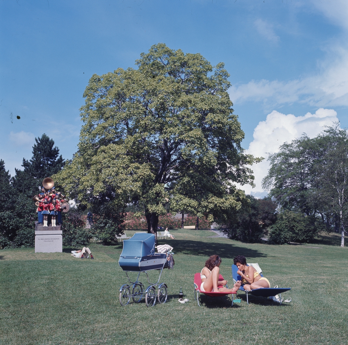 To kvinner soler seg på solsenger i parken på St.Hanshaugen, en barnevogn står ved siden av dem, i juli 1970. I bakgrunnen sees skulpturen "Musikanter" av Arne Vinje Gunderud, oppført i 1970.
