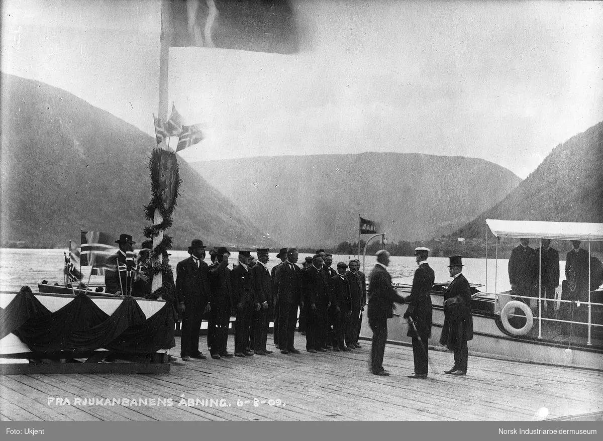 Besøk av Hans Majestet Kong Haakon under Rjukanbanens åpning. Kongen håndhilser med en mann på bryggen, ferge ligger i bakgrunnen. Mennesker stående på rekke på bryggen som er pyntet med flagg og girljander. Norges riksvåpen henger på flaggstang.