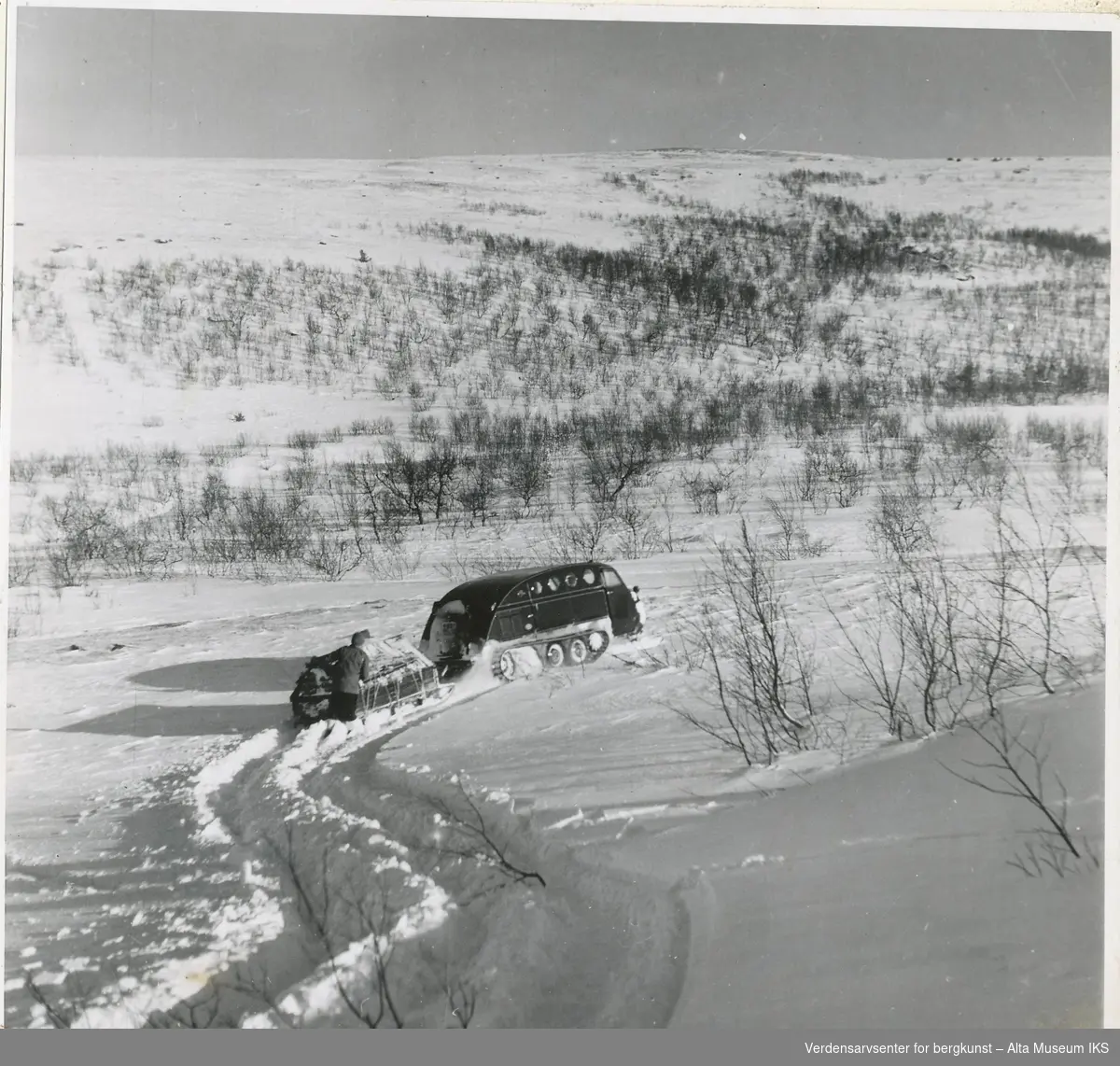 Transportproblemer i fjellet. Muligens en Bomardier 1949 B12 som trekker på en slede, men står fast, en mann står bak og dytter.