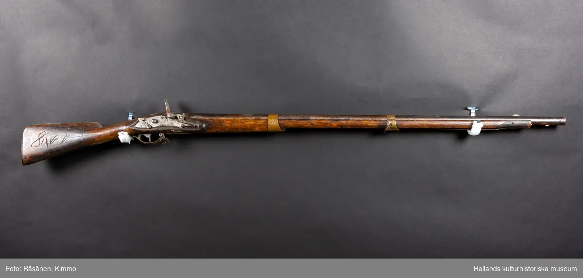 a) Gevär använt i Skarpskytterörelsen. Frontladdat med pipa och skjutanord-ning av järn. Kolv och stag under pipan av trä. Kolven järnförstärkt baktill. Två mässingsklamrar håller ihop pipa och trästag, varav ett löst. Mässingsskydd runt avtryckaren. Fästen för band att bära geväret med, men bandet saknas. Längst fram vid siktet saknas en mässingsklammer. Stämplat i kolvjärnet: "61" och "14". Ristat i träkolven: "IE (S)", "58 (12)" och "81X" samt två pilar. Pipan är rostig.b) Bajonett som går att fästa framtill på geväret, helt av järn. Vinklad framåt. Mått: längd 70 cm.