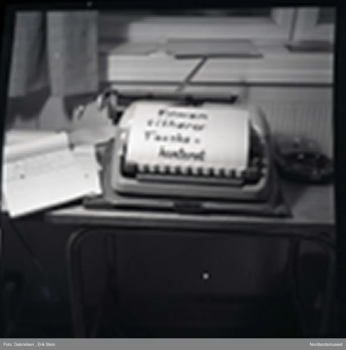 Fotografi av en gammel skrivemaskin 
som står på en pult inne i et kontor under en vindu med noen gardiner som det ligger en lapp på som står filmen tilhører 
Fauskekontoret og det ligger noen papirblokker
vedsiden av skrivemaskinen og man ser noen
mapper som ligger på vinduskarmen i kontoret 
og man ser en ovn under pulten i kontoret