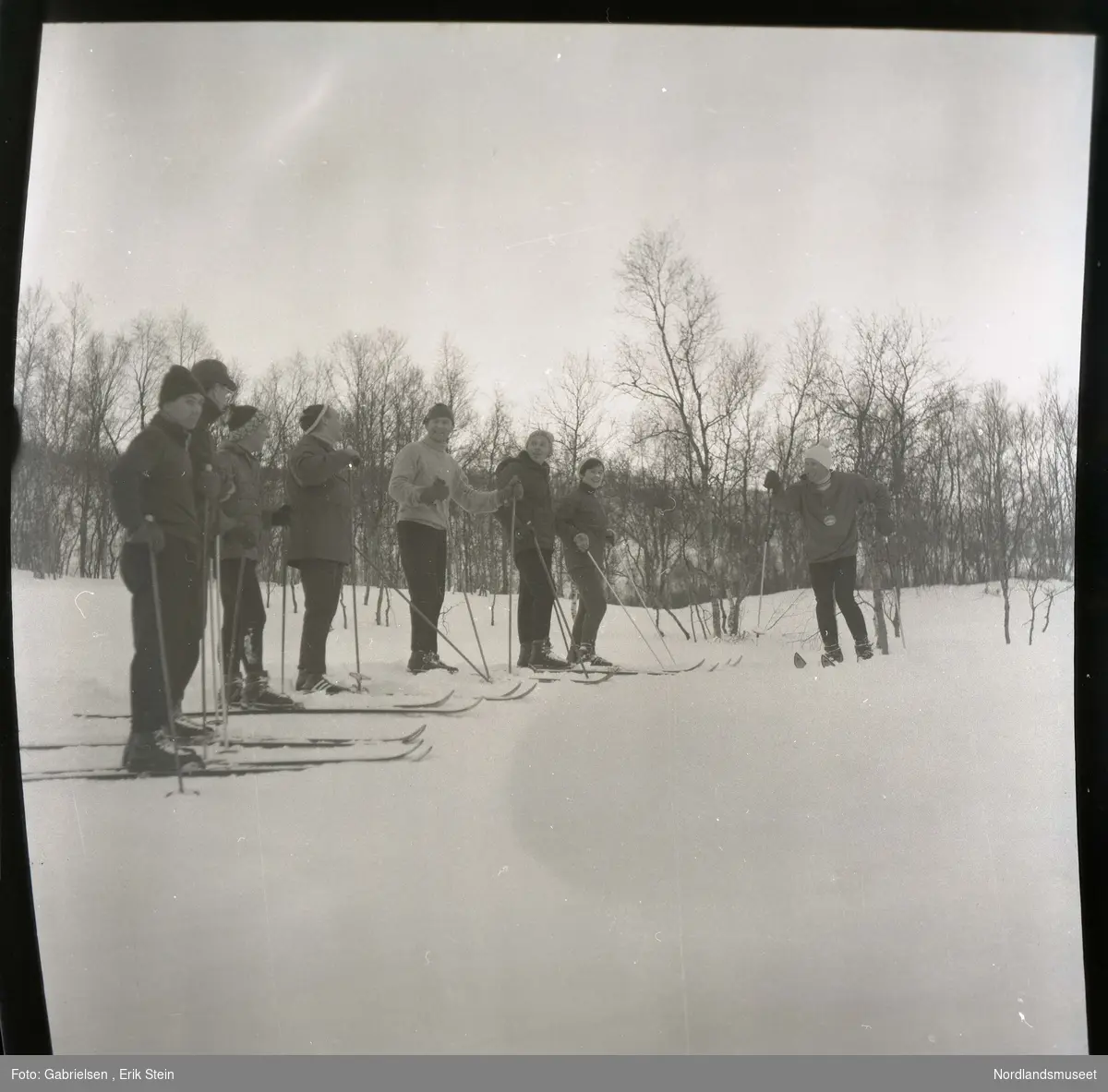 Fotografi av mange menn og kvinner med vinterklær
og ski på seg som står i snøslette vedsiden av en skog som det vokser noen trær og holder noen skistaver mens
de snakker og ler med hverandre 
og man ser et kvinnen med ski på seg 
lengst til høyre har en medalje på seg