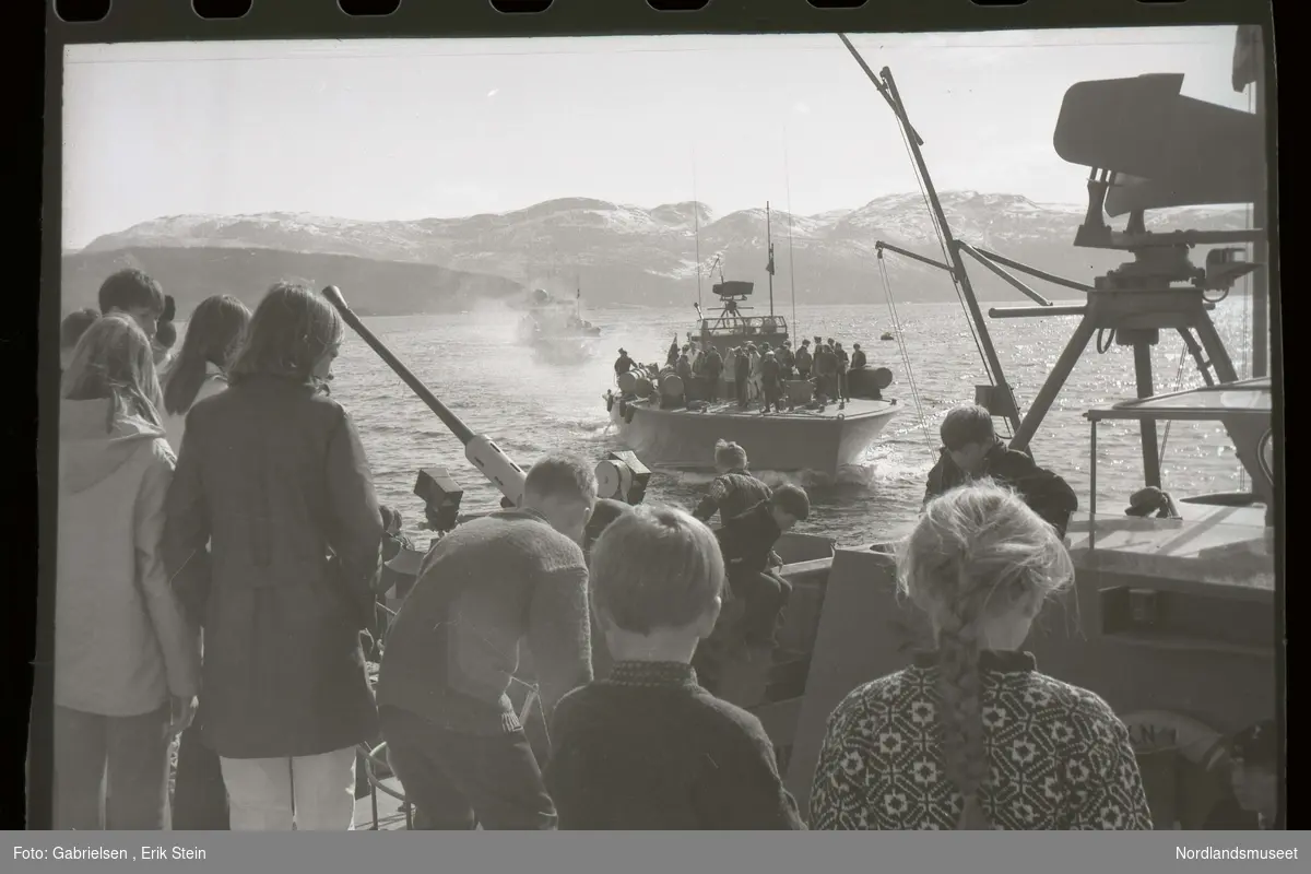 Fotografi av mange barn som står på en krigsbåt 
ute på Fauskefjorden og ser på to krigsbåter 
med mange personer på som kjører inn mot krigsbåten
som barnene står i og man ser noen anti luftvåpen
på krigsbåtene og noen kraner og antenner 
og man ser litt av Lundhalvøyen og noen fjell 
som det ligger snø på på andre siden av fjorden
