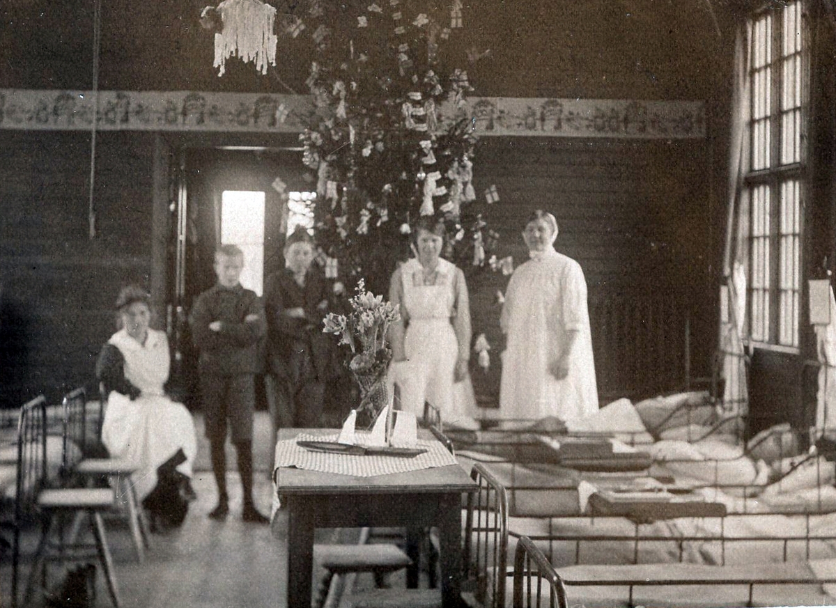 Patienter och sjuksystrar i julpyntad sjuksal i en av Kustsanatoriets träpaviljonger. En rikt pyntad och hög gran står i centrum, en lång bonad pryder väggenarna och på bordet står en julbukett med tulpaner.