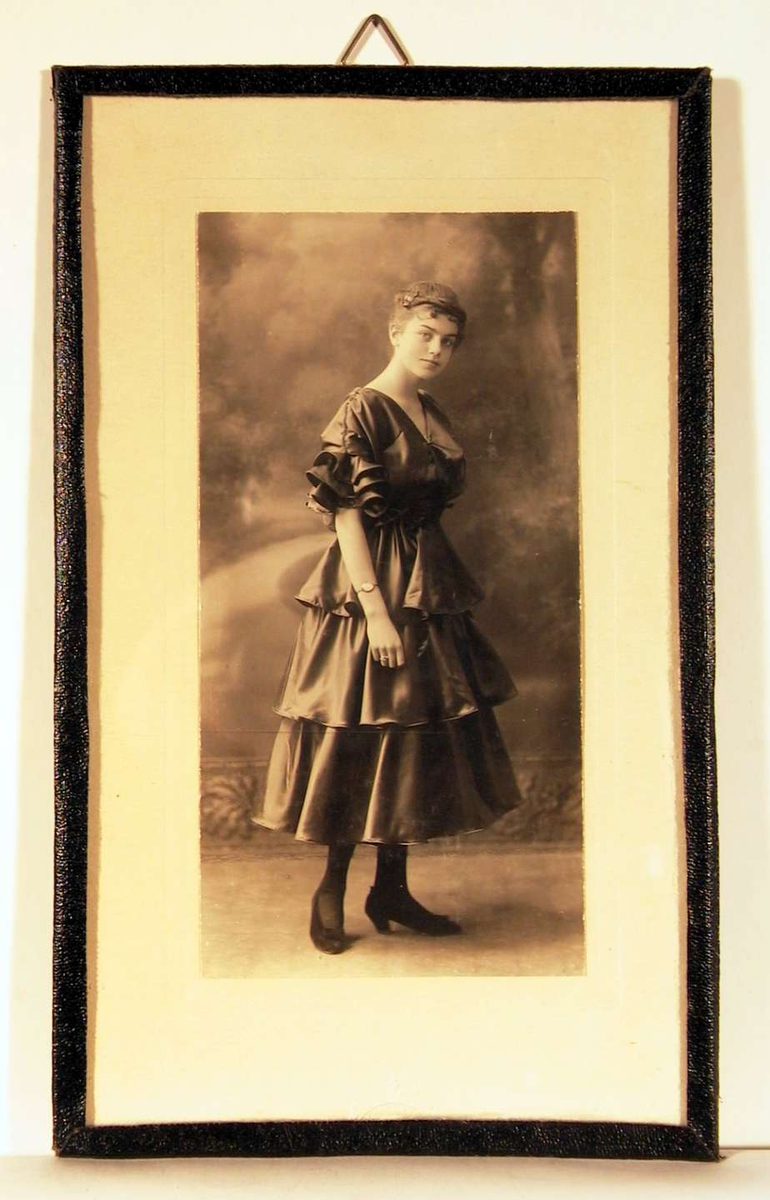 Atelierfotografi; helbilde av en ung dame i halvlang ballkjole med brede fleser i taft, sko med sløyfe og oppsatt frisyre. Bakgrunnsteppe viser en parksti kantet med trer og busker i solskinn.