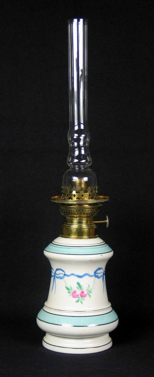 Parafinlampe i hvit keramikk med tre parafinglass. Den originale kuppelen mangler. 
Lampefoten er rund. Lampen er dekorert med to grønne border. Mellom bordene er det en blå sløyfebord med hengende blomster .