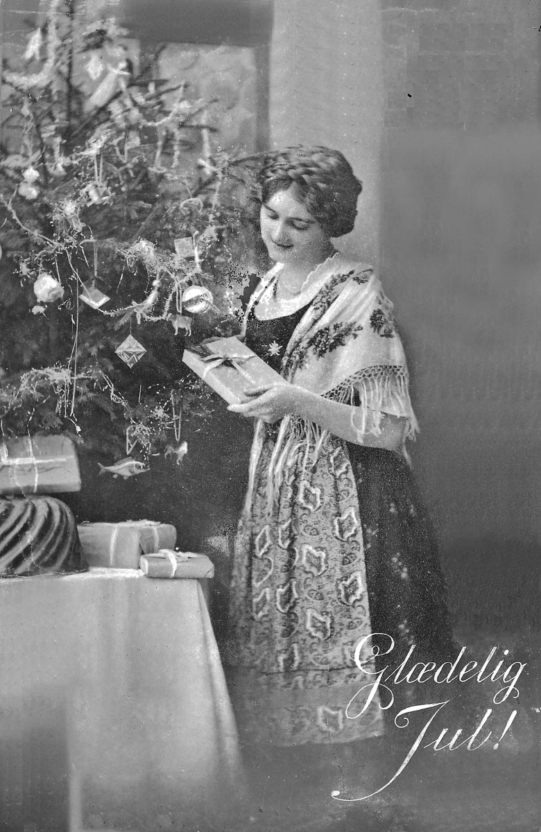 Postkort, julekort, kvinne med pakke i hendene og står ved juletre