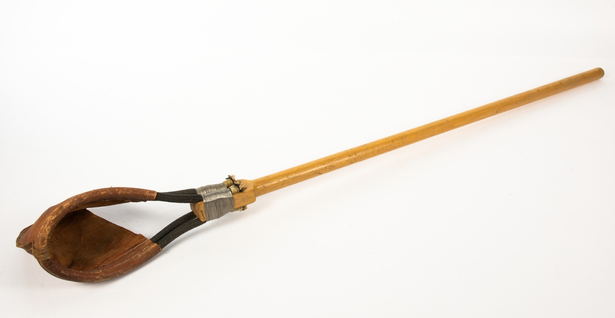 Startslunga tillverkad av trä och läder. I änden av en käpp är en "läderpåse" fäst med metalltråd.