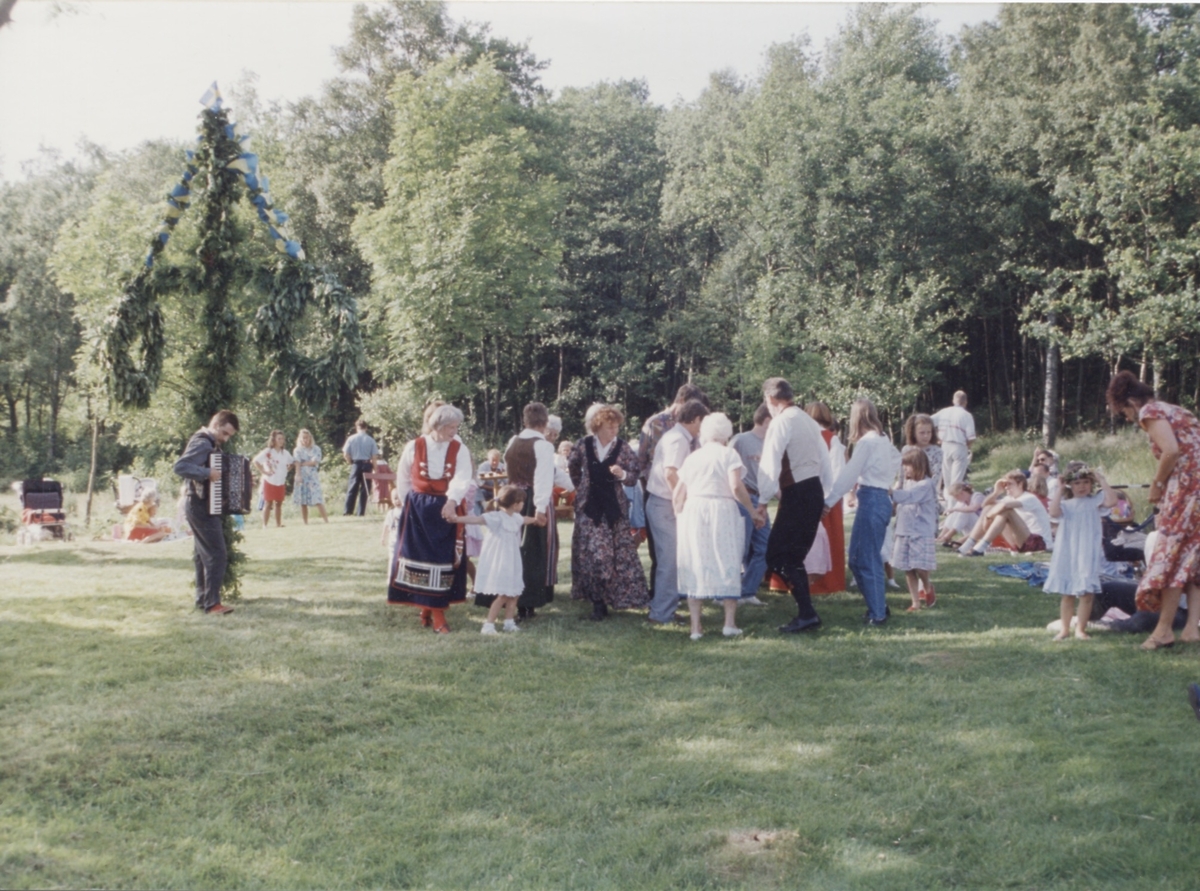 Dans kring midsommarstången vid Långåker 1:3 Hembygdsgården 1993. Till vänster i Värendsdräkt ses Marianne Jansson (1924-2006). Den manlige okände dragspelaren är från Göteborg. 
Relaterade motiv: A03389 - A03408.