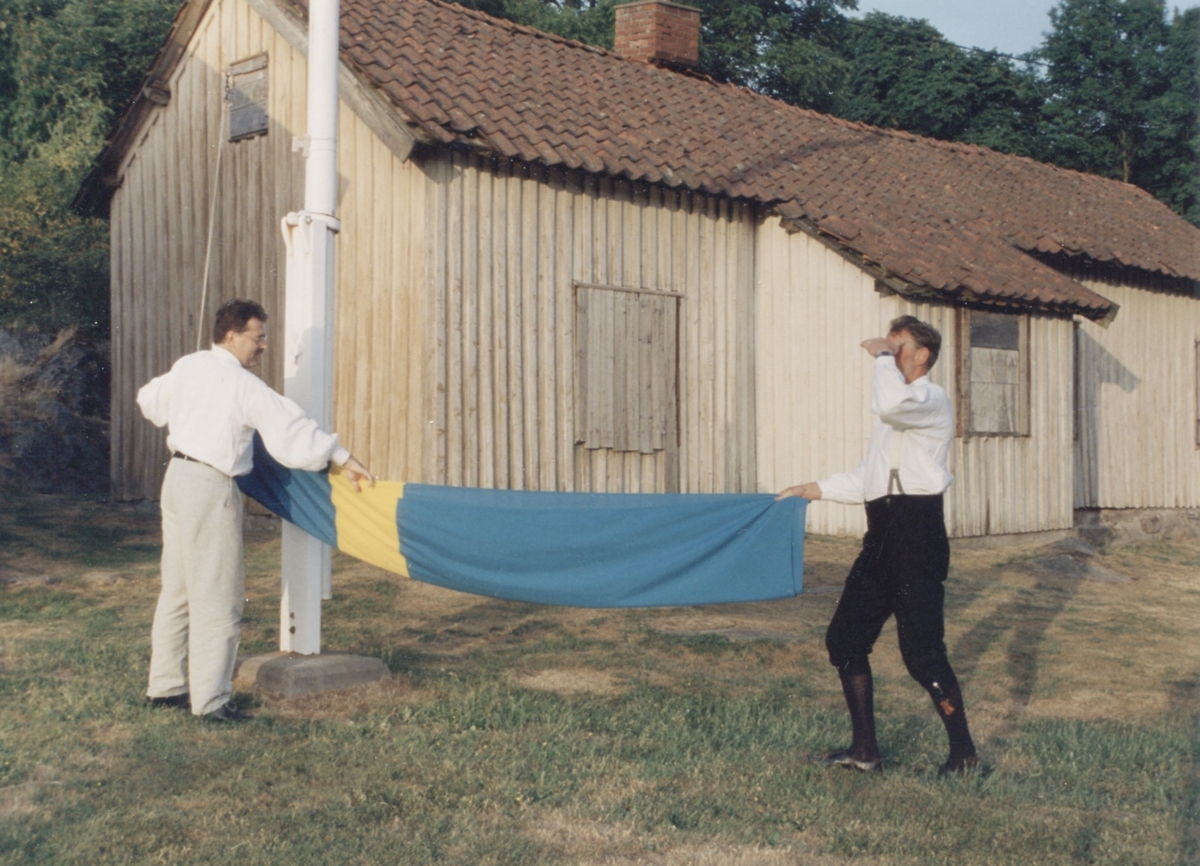 Staffan Bjerrhede och Berny Gustafsson viker ihop svenska flaggan efter midsommarfirande vid Hembygdsgården Långåker 1:3 (ej i bild) 1993. I bakgrunden ses huset Långåker 1:2 "Davids", "Derras".
Relaterade motiv: A03389 - A03408.
