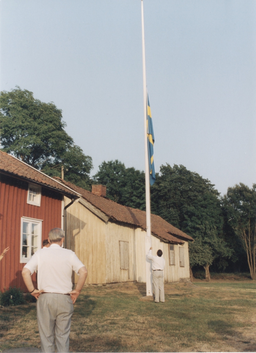 Staffan Bjerrhede halar ner flaggan efter hembygdsföreningens midsommarfirande 1993. Berny Gustafsson och okänd man ser på. Till vänster ses den röda Hembygdsgården Långåker 1:3 och i bakgrunden Långåker 1:2 "Davids", "Derras".
Relaterade motiv: A03389 - A03408.
