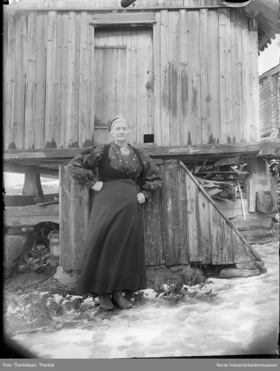 Kvinne i stakk og liv med sølje i halsen stående foran bur på den gamle husmannsplassen Merde under Nordre Tinnes.