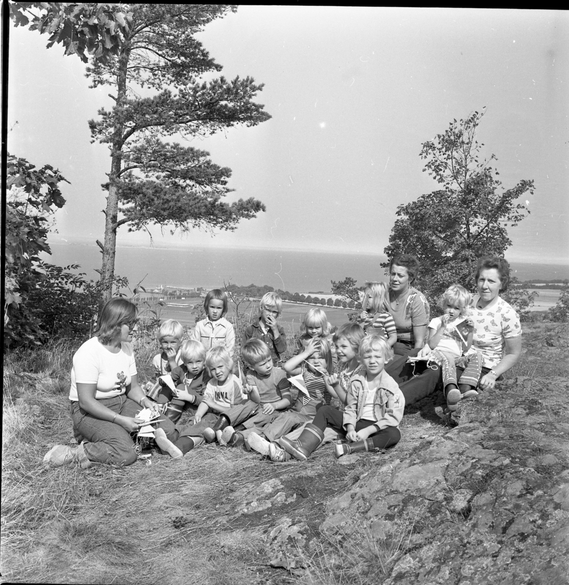 Vuxna och barn sitter på Grännaberget med utsikt mot Gränna hamn. Det är sommar.