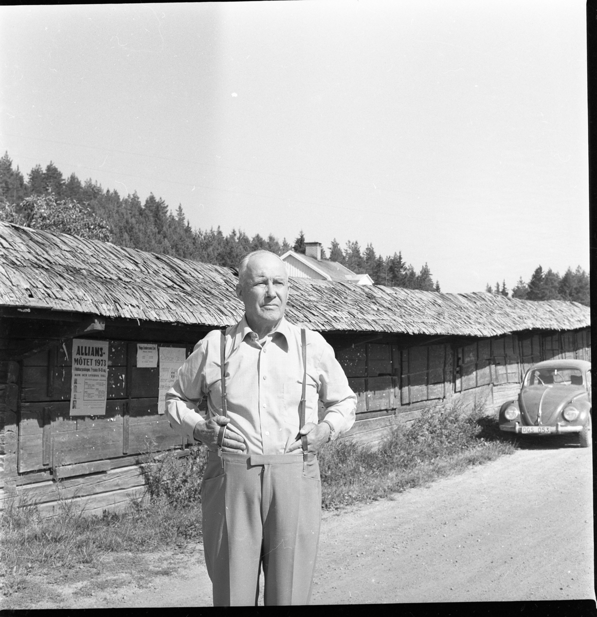 En man i ljus skjorta och byxor med hängslen står vid marknadsbodar i Adelöv. En VW-bil står i bakgrunden.