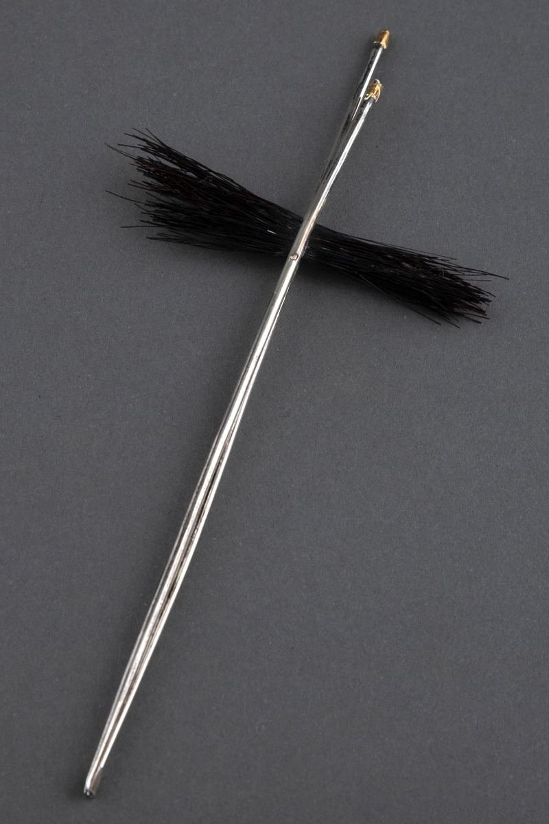Tre nåler som består av to blanke metallpinner av ulik lengde som er festet sammen. På tvers, mellom dem, er det en bunt svart hestetagl. Pinnene er forgylt på toppen.