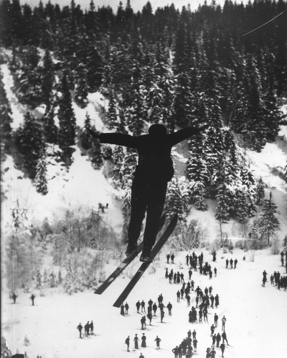 I fritt svev i hoppbakken med mange tilskuere tilstede. Fotografert  7. februar 1909.