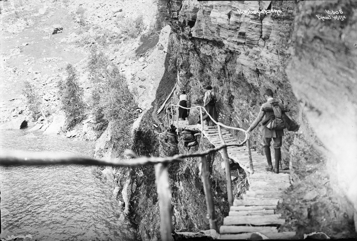 Tre fjellvandrere forserer Nesbøgalden (gald sies å ha betydningen 'vanskelig framkommelig plass') på treplattinger med rekkverk innfelt i det loddrette berget, 1914.