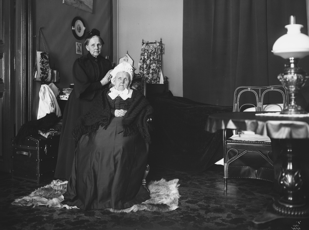 Bispinne Grimelund sittende på en stol i soverommet, med datteren stående ved siden av. Fotografert 1909.