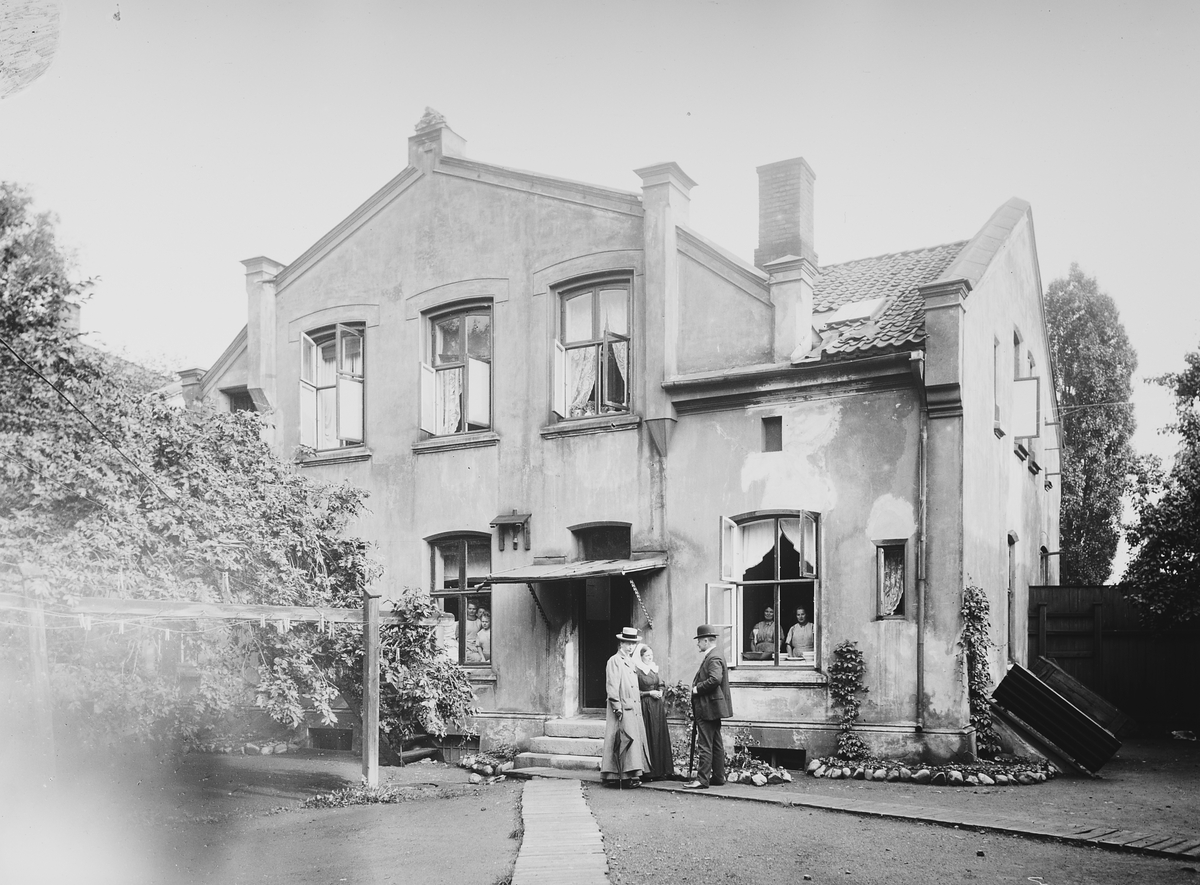 To kvinner og en mann, presten Oscar Hvalbye, foran Indremisjonens gamlehjem. To ansatte i vinduet. Fotografert 1911.