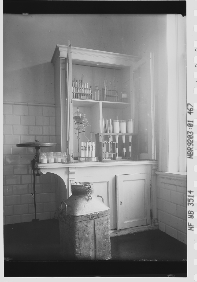 Skap med prøver av melk fra De Forenede Meierier. Foran står en stor melkespann. Fotografert 1911.
