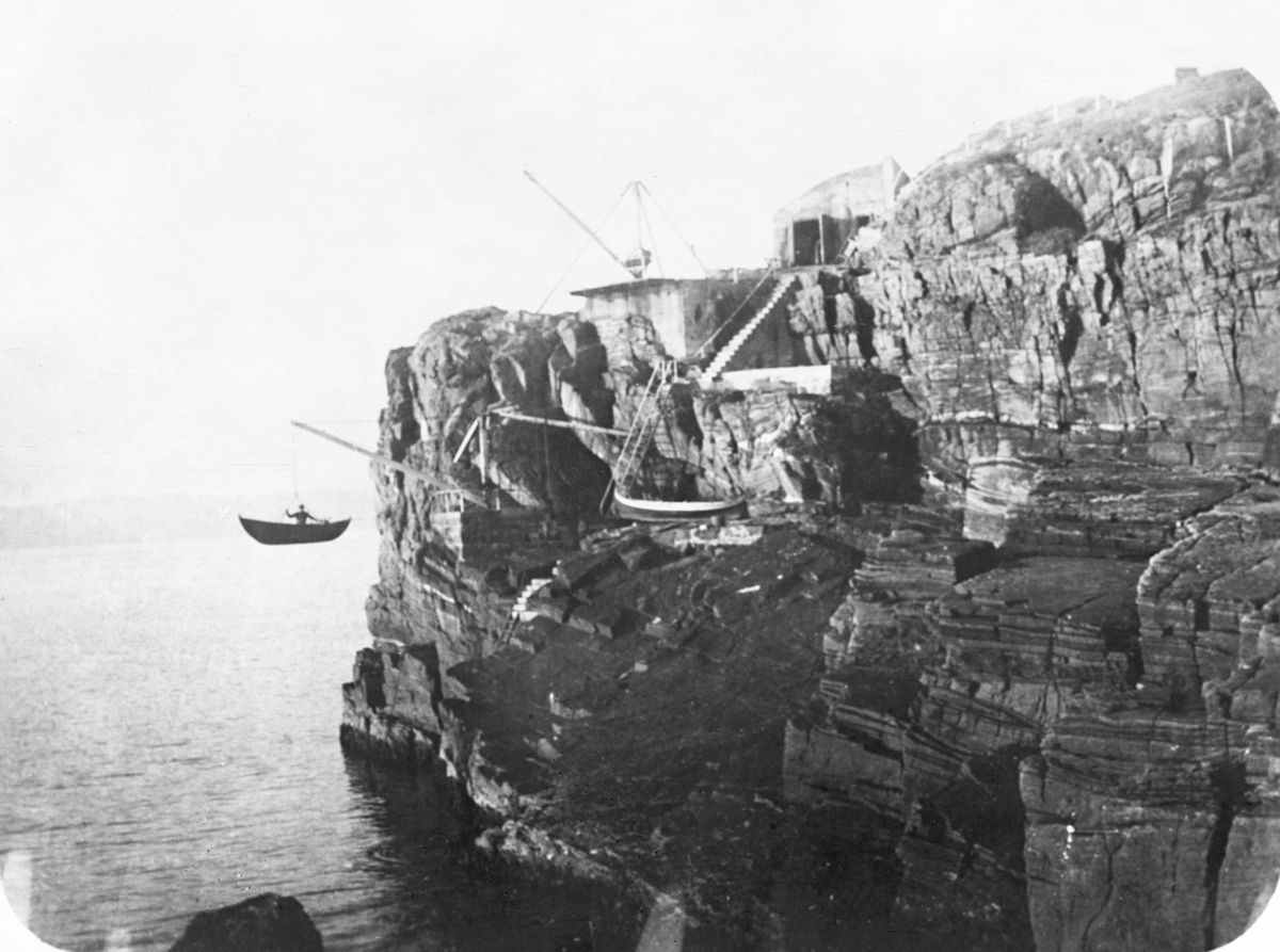 Mann i båt låres i en båt ned fra fjell mot sjøen. Trapper går ned fjellvegg, båt ligger lagret i fjellet. Svinøy fyr, Herøy. Fotografert 1923.