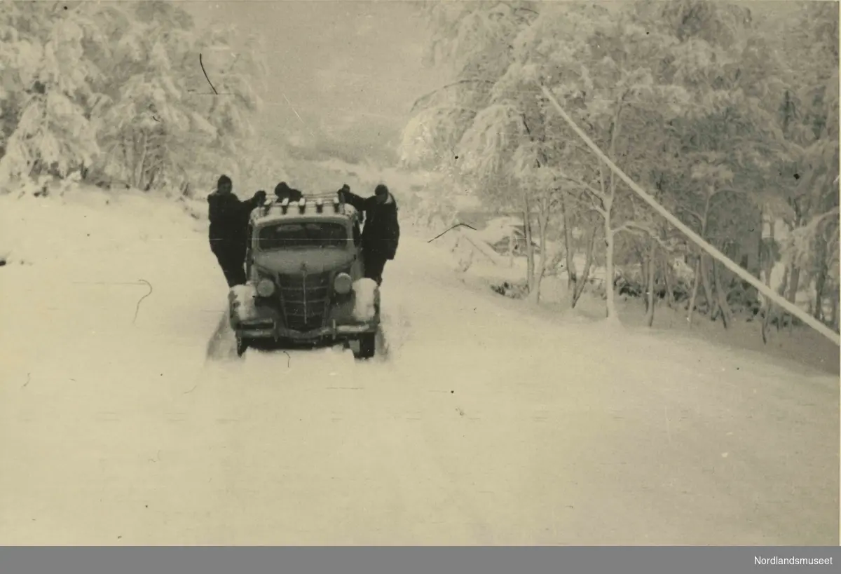 En bil står eller kjører på en snødekt vei. Tre personer utenfor.