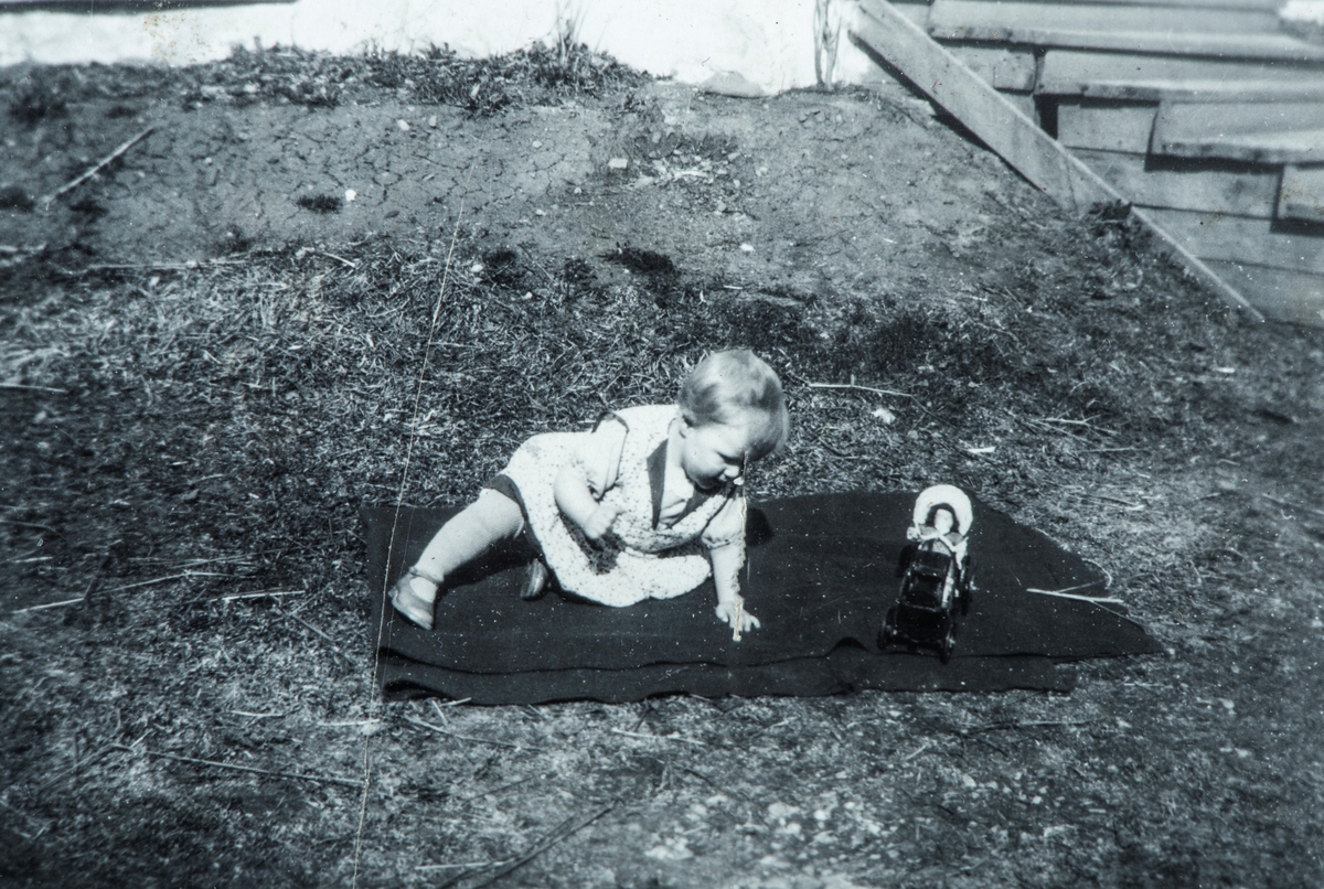 Øvre Tomter (60/1). Ragnhild Prestrud sitter ute å leker på et teppe på grasbakken foran hovedbygningen. 1947.