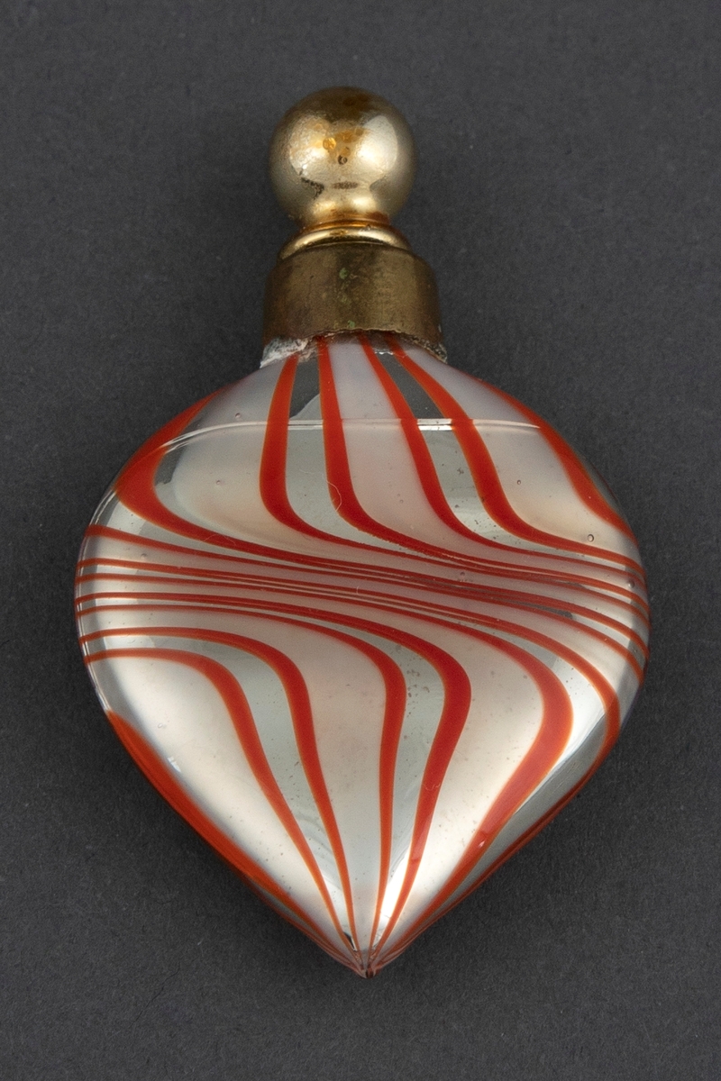 Flat hjerteformet flakong i melkehvit opak glass med vridd stripemønster i rødt og sølv. Flakongen har en hals av metall og en gullfarget pipette med kuleformet lokknapp på toppen.