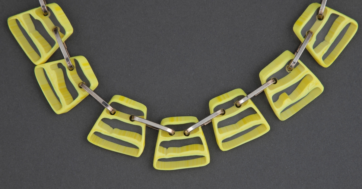 Halssmykke med store, trapesformede glassbiter i gult og klart glass, der klare og gule horisontale striper er omkranset av gult. Glassbitene er hengslet sammen med grove sølvringer.