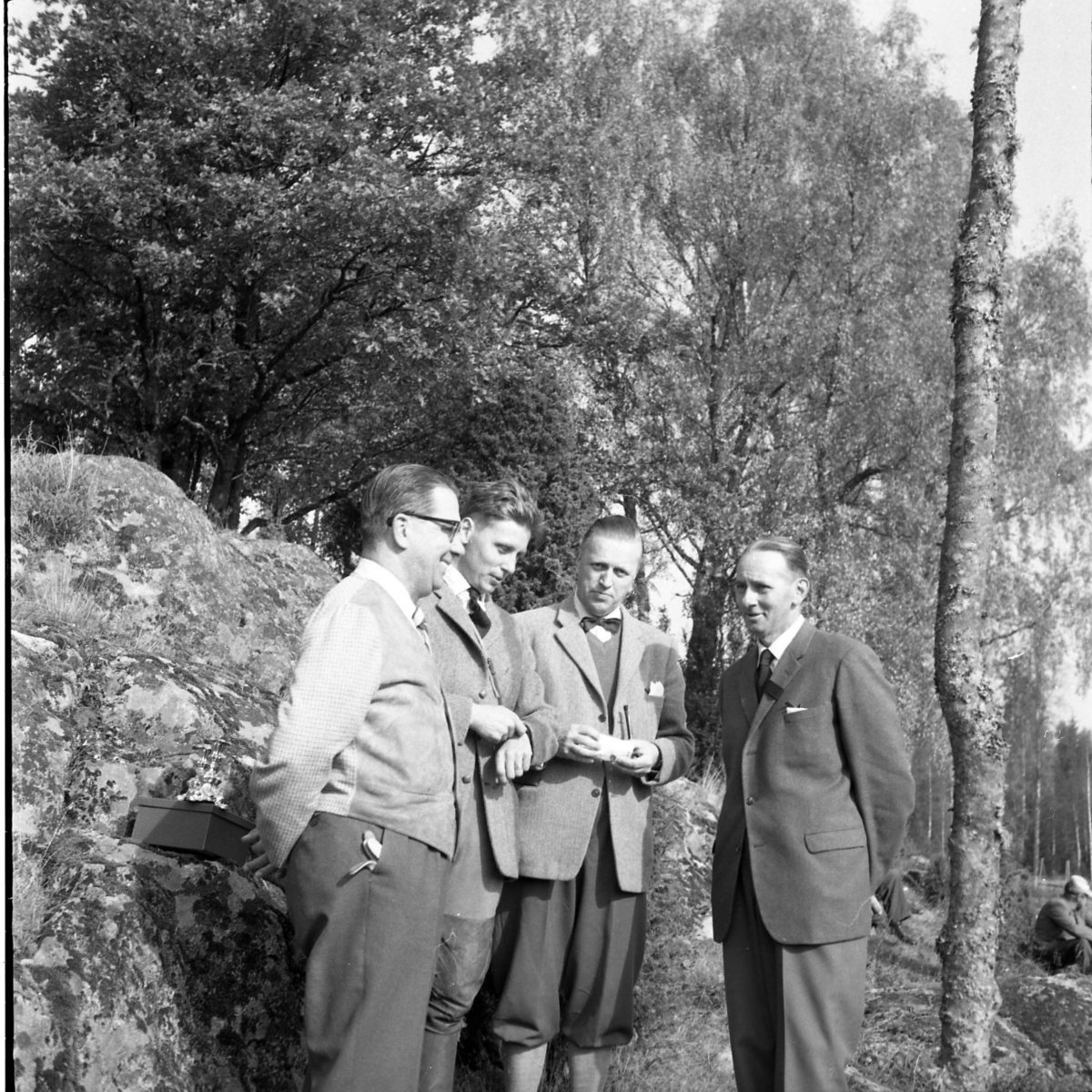 Fyra män står axel mot axel och pratar. De bär kavaj och slips. En man håller på att stoppa en pipa.