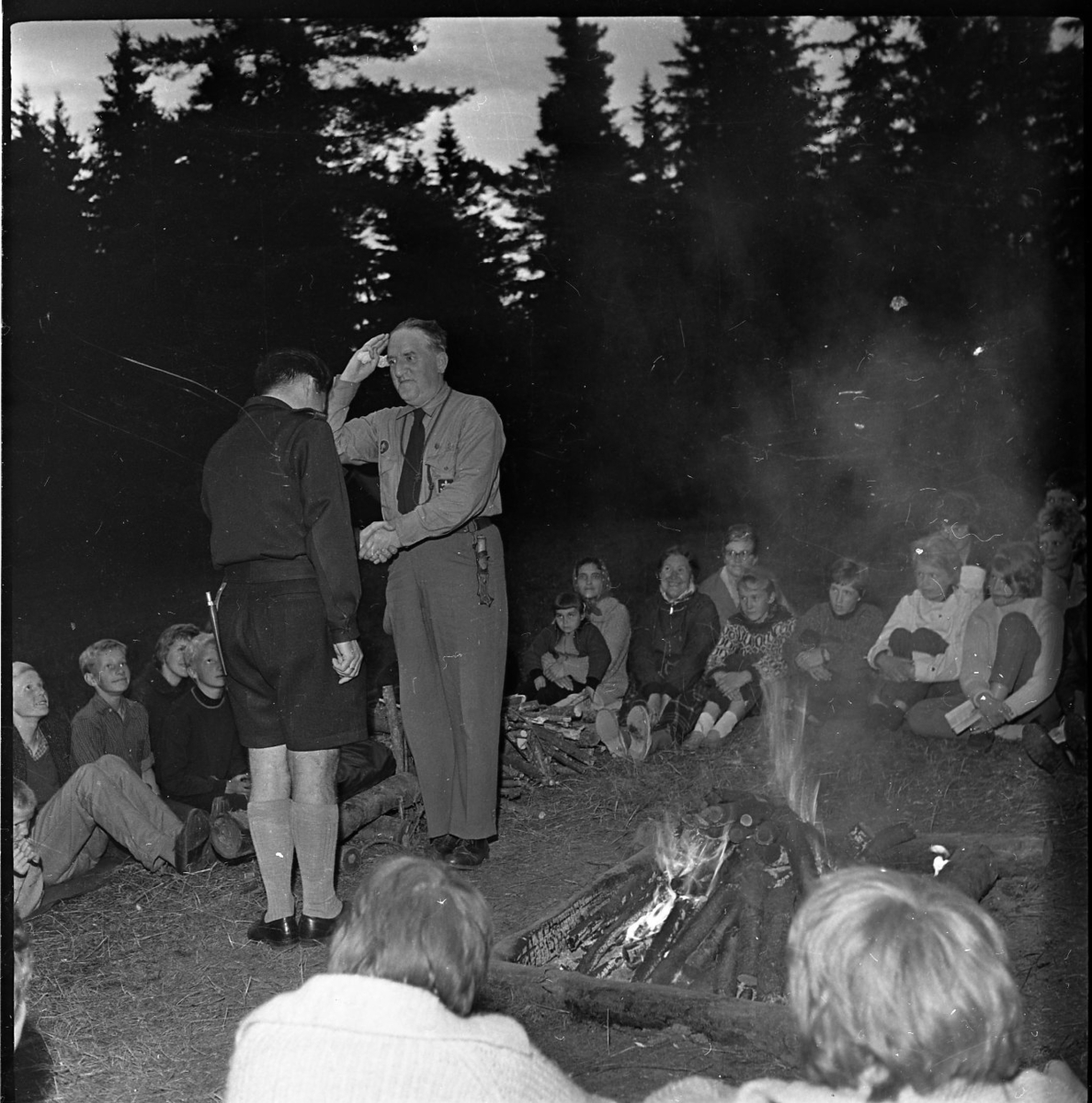 Ett gäng ungdomar sitter runt en lägereld. Två  scoutledare står upp. De skakar hand och en av dem gör honnör.