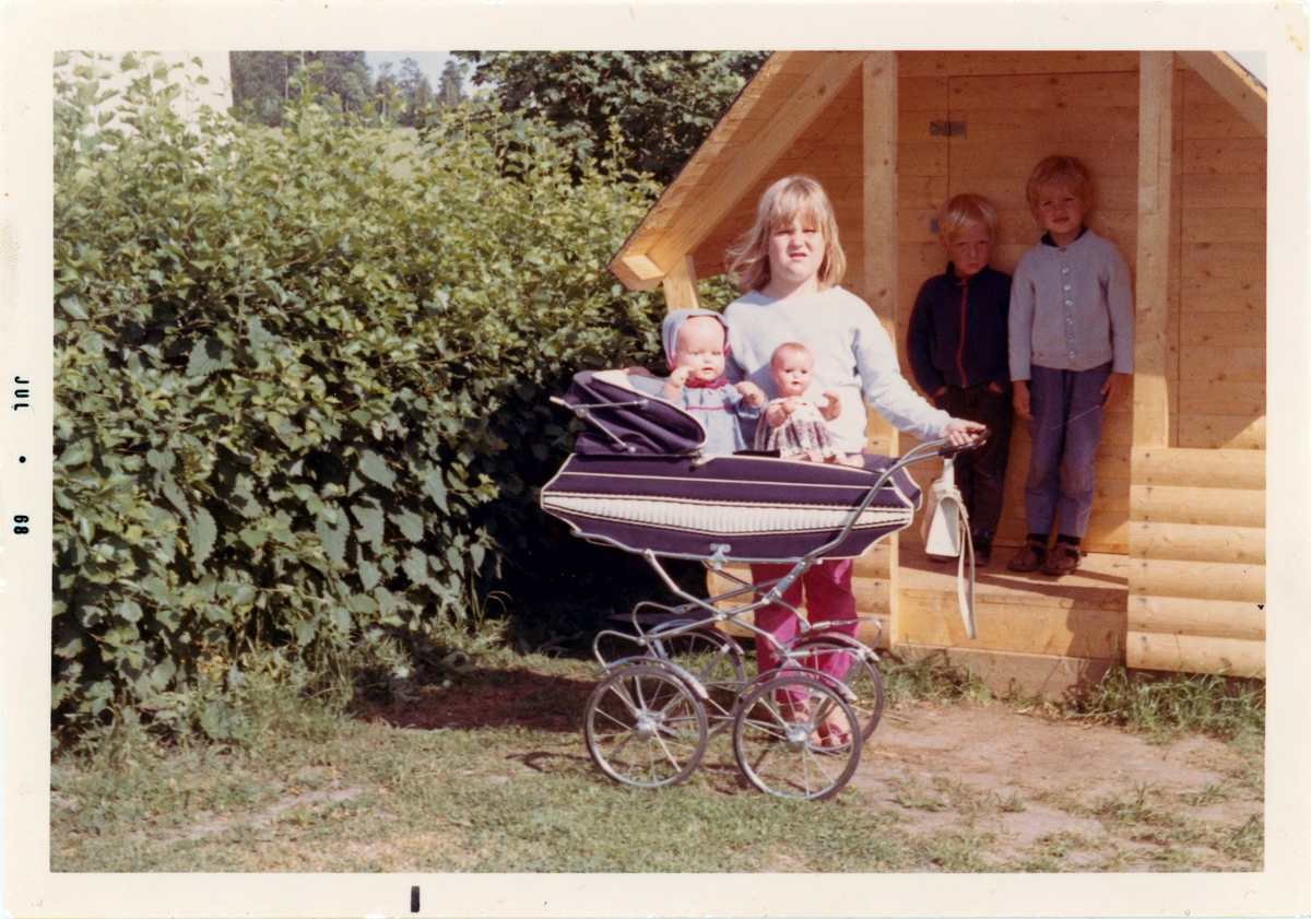 Tre barn och en dockvagn med dockor framför Erika Karlssons lekstuga på Stensholm i Huskvarna sommaren 1968. Dockvagnen och den vänstra dockan, Carina, tillhör Erika, som inte är med på bilden.