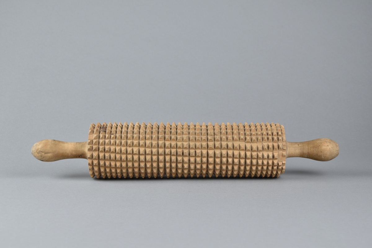 Brödkavel tillverkad i trä. Cylinderformad med handtag på var sida, svarvad i ett stycke. Kaveln med inskurna täta taggar. 