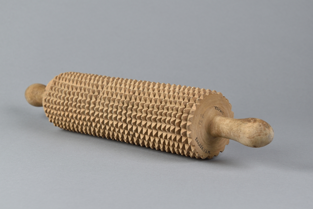 Brödkavel tillverkad i trä. Cylinderformad med handtag på var sida, svarvad i ett stycke. Kaveln med inskurna täta taggar. 