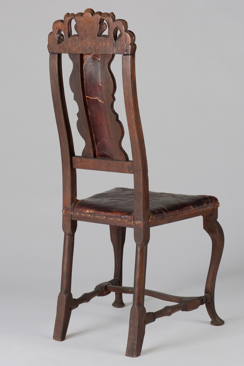 Høyrygget barokkstol med skinnsete og skinntrukket ryggfjøl. Stolen har et toppstykke med kronet akantus. Stolbenene foran er svungne og de bakerste er rette, benene er satt i et H-kryss.