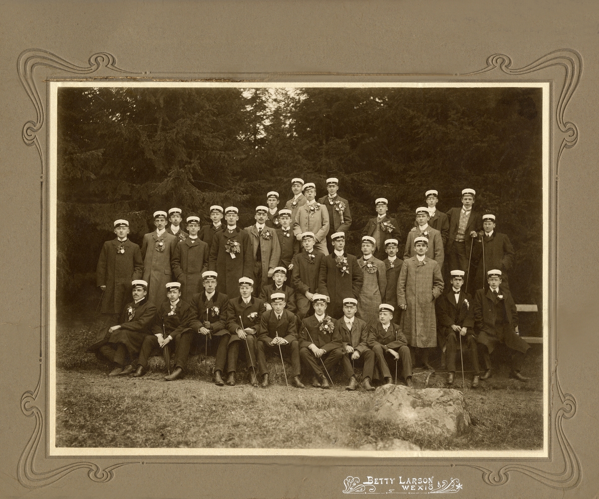 1907 års studenter i Växjö har samlats för ett gruppfoto utomhus.