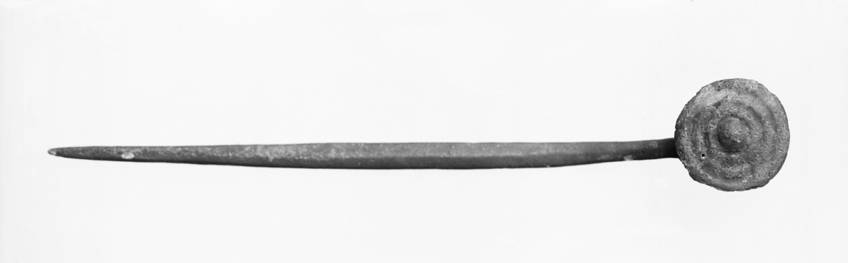 Naal af bronce af formen MÜLLER, fig. 301, hvis øvre del er noget bøiet og bærer en lodretstaaende, rund bronceplade; pladen er godt 2 cm. i tvermaal, og dens forside er orneret med tre fordybede cirkler samt i midten en liden halvrund forhøining. Naalens samlede længde er 13,1 cm. (fig. 9)
Revisjon 2022: Nål i kobberlegering med plate, Baudous type XXV B 2b. Den øverste delen av nålen er bøyd, der en loddrett flat rund plate er festet.  Platen har dekor i form av tre konsentriske sirkler med en rund forhøyning i midten.