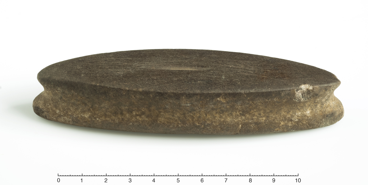 Beltestein av rødbrun kvartsitt, av den slanke form R. 155. Steinen har innfatningsfure og merker av slit på oversiden. Lengde 13,3 cm.