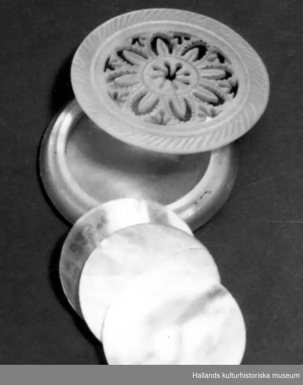 Rund ask (a) med lock (b) av pärlemor. Locket är perforerat i form av en stjärna. Asken innehåller tre marker (c, d, e) också av pärlemor. Markerna är runda och släta. 