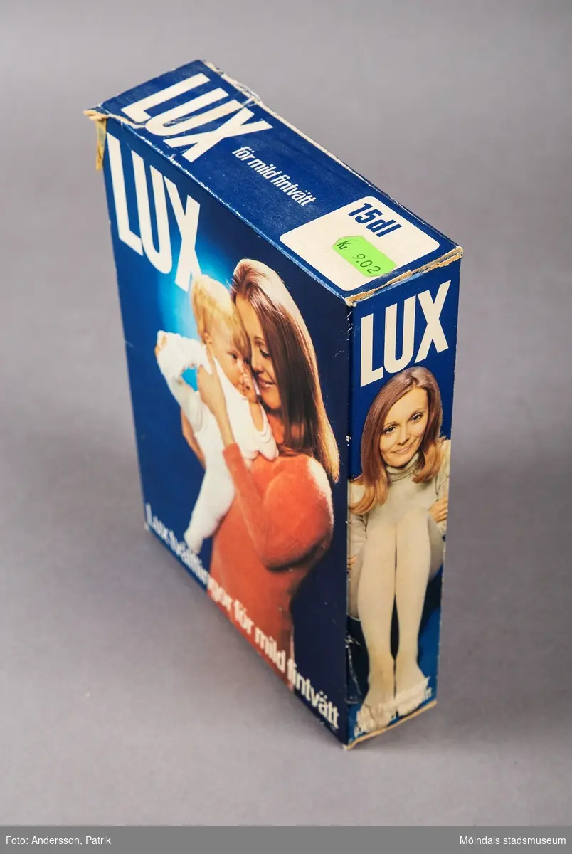 En rektangulär djupblå kartong som tidigare innehållit tvättmedel. På kartongens övre framsida står det Lux med stora vita versaler. Paketets framsida domineras av en bild på en kvinna iförd en korallröd långärmad klänning. Kvinnan ler och håller en vitklädd bebis i famnen. Bebin har tummen i munnen och sträcker ut sitt högra ben. Bilden har en intressant färgsättning och utsrålar i sin helhet harmomi och mjukhet. Texten nedtill på kartongen lyder: Lux tvålflingor för mild fintvätt.
