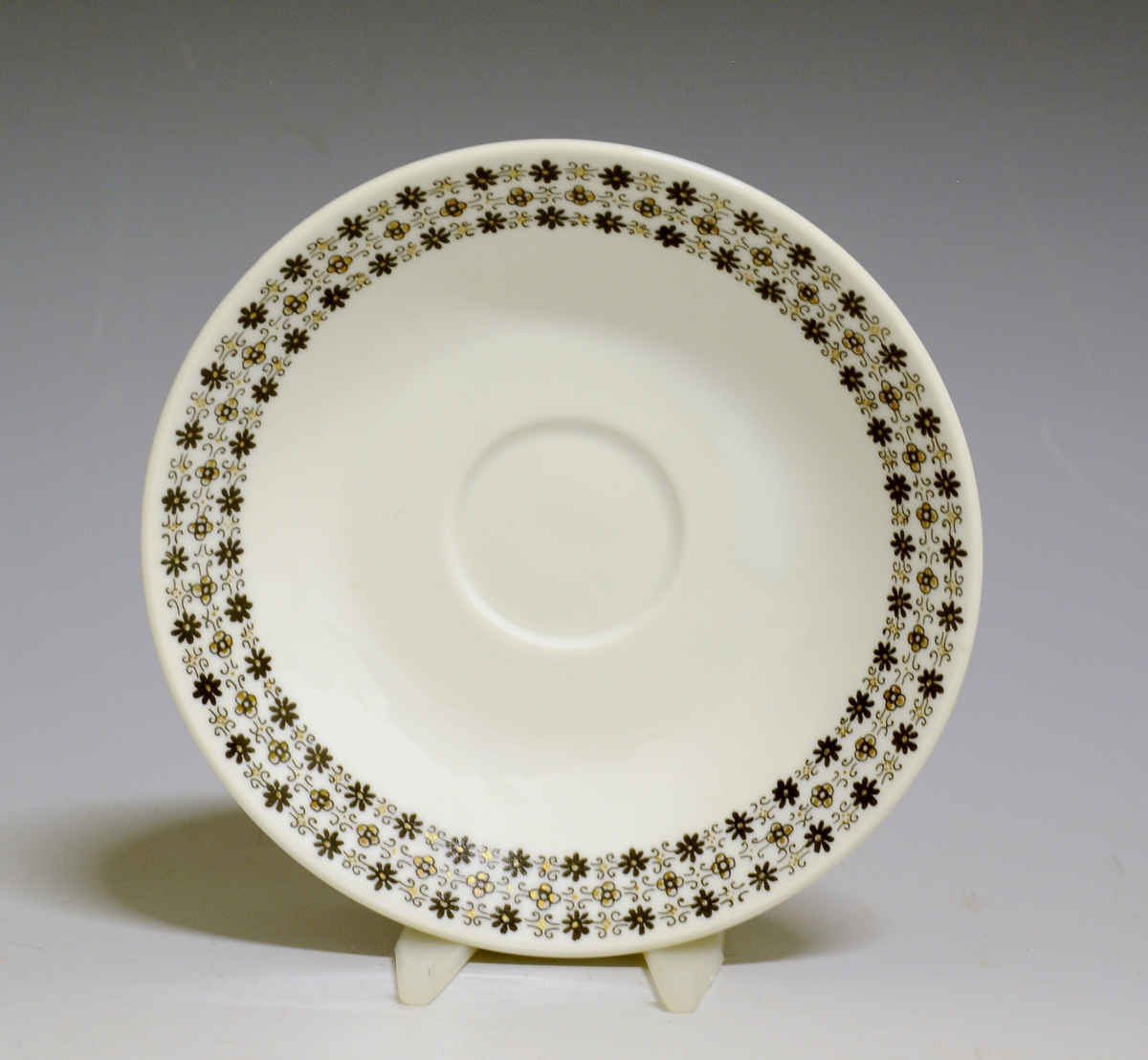 Kaffeskål av porselen med hvit glasur og bord med småblomstret trykkdekor i sort og gull langs kanten.
Modell: Petita (eller variant av)

