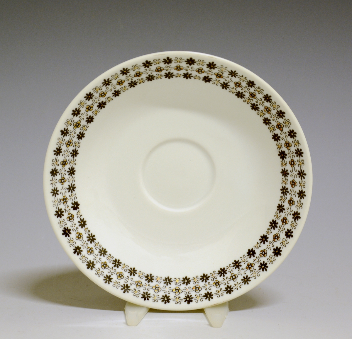 Kaffeskål av porselen med hvit glasur og bord med småblomstret trykkdekor i sort og gull langs kanten.
Modell: Petita (eller variant av)
