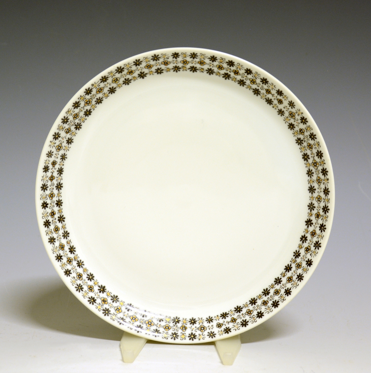 Asjett av porselen med hvit glasur og bord med småblomstret trykkdekor i sort og gull langs kanten.
Modell: Petita (eller variant av)