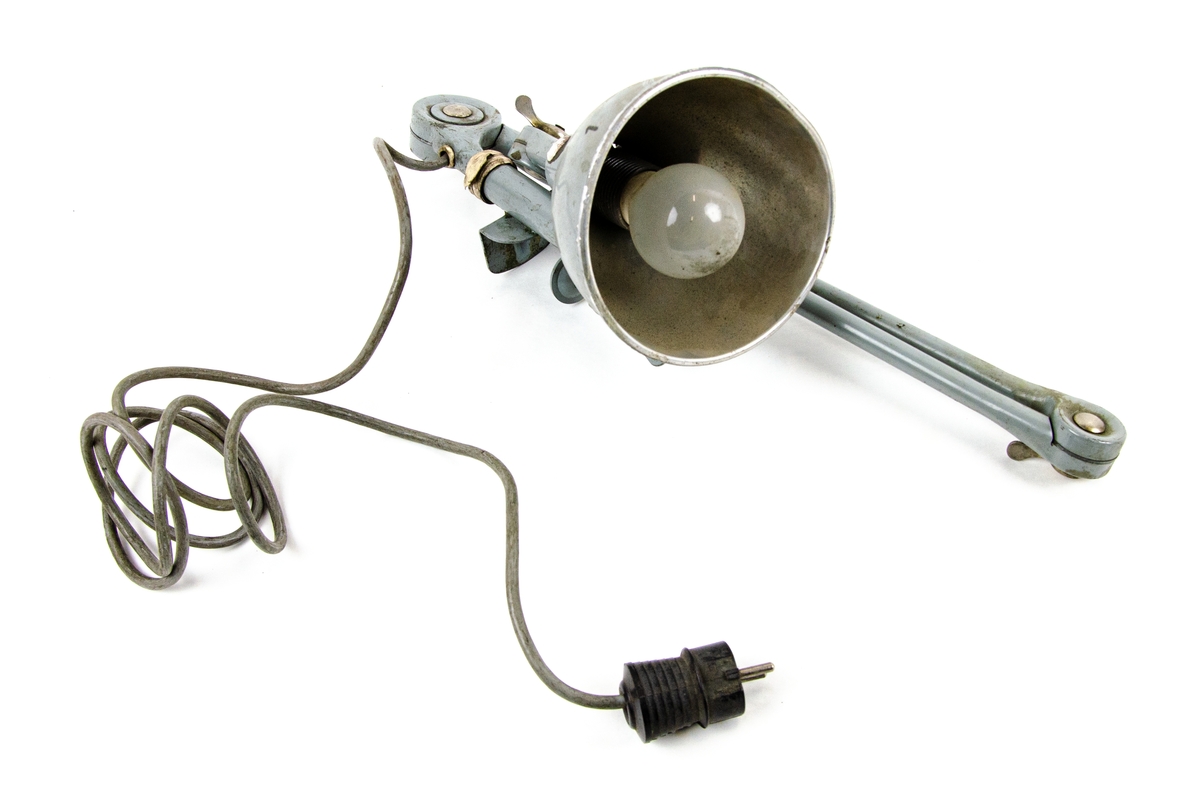 Bordslampa i grå metall, tre roterande delar och skruvklämma för att fästa lampan på ett bord och eluttag.