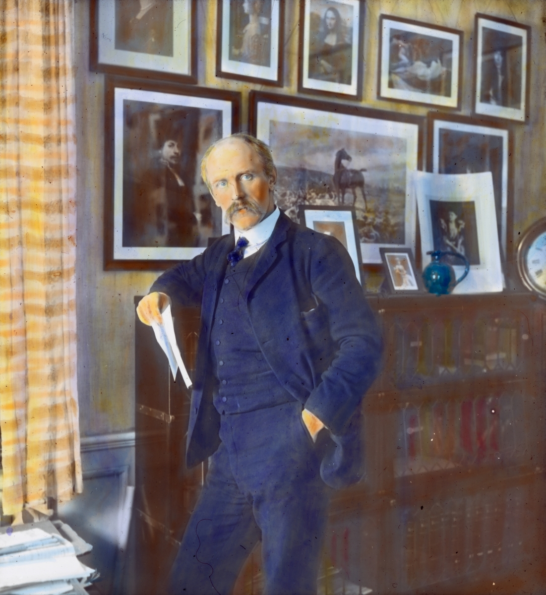Håndkolorert dias. Portrett i helfigur av polarforskeren Fridtjof Nansen. Nansen lener seg mot en bokhylle og holder papirer i hånden. På veggen bak ham henger innrammede portretter.