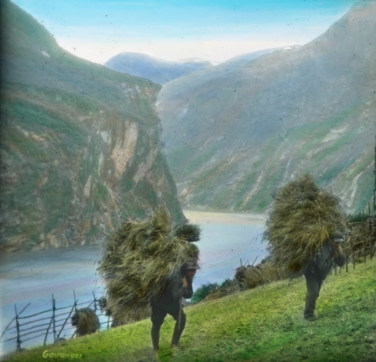 Håndkolorert dias. To menn bærer høy opp en bratt fjellside i Geirange. Bak står hersjene fulle av høy.