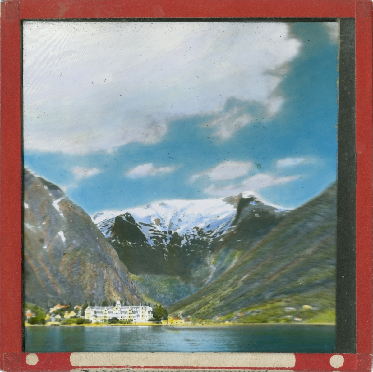 Håndkolorert lysbilde. Bilde av Kviknes hotell i grønt landskap. I bakgrunnen ses fjell med snø.
